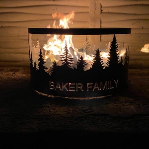 Op de afbeelding: Een zwarte metalen vuurkorf met een silhouet van een bos en de tekst "BAKER FAMILY". De vuurkorf is aangestoken met vlammen erin.