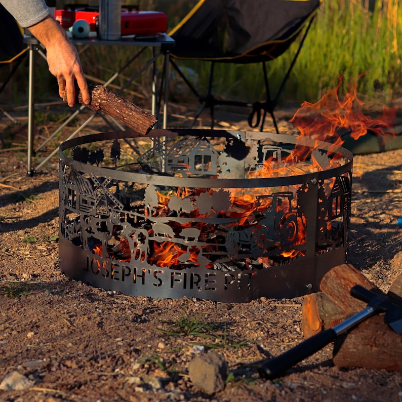 K&ouml;nnte beinhalten: Ein schwarzer Metall-Feuerplatz mit einem ausgeschnittenen Design einer Farmszene. Der Feuerplatz ist von Flammen umgeben und tr&auml;gt die Aufschrift "Joseph's Fire Pit" auf der Vorderseite.