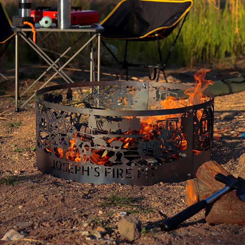 K&ouml;nnte beinhalten: Ein schwarzer Metall-Feuerplatz mit einem ausgeschnittenen Design aus B&auml;umen, H&auml;usern und einem Traktor. Der Feuerplatz brennt mit Flammen und tr&auml;gt die Aufschrift "Joseph's Fire Pit" auf der Vorderseite.