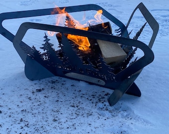 May include: A black metal fire pit with a cut-out design of trees and the words "Gather 'round the Fire Pit". The fire pit is sitting on a snowy surface with flames burning inside.