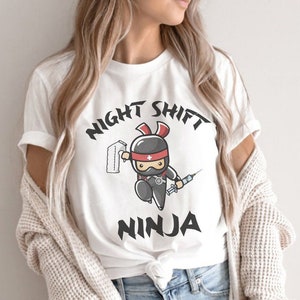 May include: White t-shirt featuring a cartoon ninja wearing a medical mask and holding a syringe and a roll of gauze. The text "Night Shift Ninja" is printed in black above the graphic.