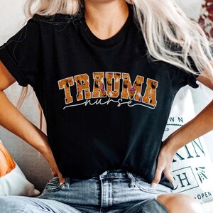 Puede incluir: Una camiseta negra con el texto "Trauma Nurse" en letras naranja y negras. Las letras est&aacute;n llenas de un patr&oacute;n de iconos de Halloween, incluyendo calabazas, murci&eacute;lagos, fantasmas y sombreros de bruja.