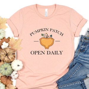 K&ouml;nnte beinhalten: Ein pinkfarbenes T-Shirt mit dem Text "Pumpkin Patch - Open Daily" und einer Grafik einer K&uuml;rbis mit Bl&auml;ttern. Das Shirt liegt auf einer wei&szlig;en Oberfl&auml;che mit Herbstdekorationen.