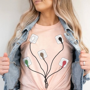 May include: A pink t-shirt with a graphic of five white electrical outlets with different colored wires connected to them. The outlets are arranged in a tree-like shape.