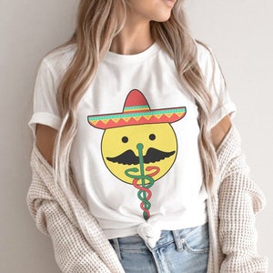 K&ouml;nnte beinhalten: Ein wei&szlig;es T-Shirt mit einem gelben Smiley, der einen roten, gr&uuml;nen und gelben Sombrero tr&auml;gt. Der Smiley hat einen schwarzen Schnurrbart und eine gr&uuml;ne Schlange kommt aus seiner Nase.