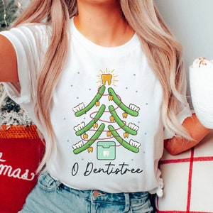 May include: White t-shirt with a green Christmas tree design made of teeth. The tree is decorated with gold floss and a gold tooth star on top. The text "O Dentistree" is printed below the tree.