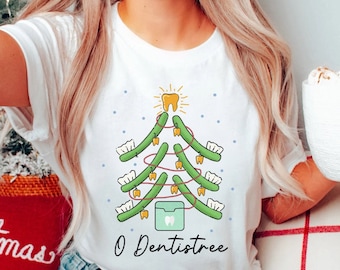 Puede incluir: Camiseta blanca con un diseño de árbol de Navidad verde hecho de dientes. El árbol está decorado con hilo dental dorado y una estrella dorada en forma de diente en la parte superior. El texto "O Dentistree" está impreso debajo del árbol.