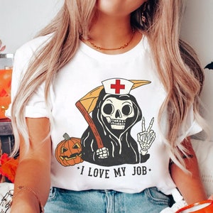 May include: White t-shirt with a graphic of a skeleton wearing a nurse's hat and holding a scythe. The skeleton is wearing a black robe and has a peace sign. The text "I love my job" is printed below the graphic.
