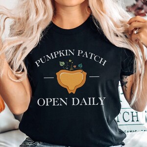 K&ouml;nnte beinhalten: Ein schwarzes T-Shirt mit dem Text "Pumpkin Patch - Open Daily" und einer Grafik einer K&uuml;rbis mit gr&uuml;nen Ranken.