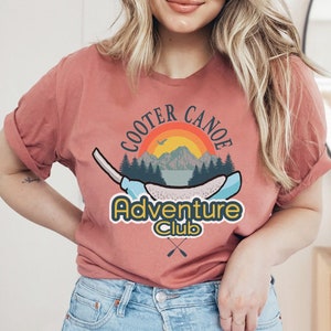 May include: A woman wearing a rust-colored t-shirt with a graphic design of a canoe, a sunset, and mountains. The text on the shirt reads "Cooter Canoe Adventure Club".