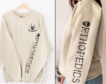 May include: A beige sweatshirt with a black skull and bones design on the sleeve and the text "ORTHOPEDICS" on the back. The design includes a hand with a rock and roll hand gesture and lightning bolts.
