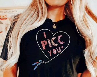 May include: Black t-shirt with a heart-shaped graphic that reads "I PICC YOU" in pink and white. The design includes a climbing rope and carabiners. The shirt is a casual wear item.