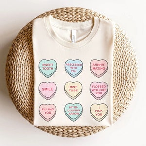 May include: A white t-shirt with nine colorful heart-shaped candy designs. Each candy has a different message, such as "Sweet Tooth", "Abscessed With You", "Ahhhh-Mazing", "Smile", "Mint To Be", "Flossed Without You", "Filling You", "Hit Bi-Cuspid Arrow", and "I Pick You".