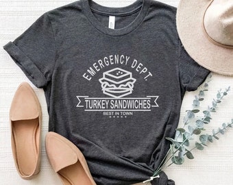 Może przedstawiać: Szary t-shirt z białym nadrukiem, na którym widnieje napis "Emergency Dept. Turkey Sandwiches Best in Town" oraz stylizowany obrazek kanapki.