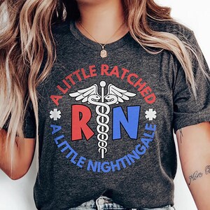 Puede incluir: Camiseta gris oscuro con el texto "A Little Ratched, A Little Nightingale" en un diseño circular. El diseño incluye las letras "RN" en rojo y azul, un símbolo médico y estrellas.