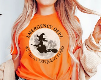 May include: Orange t-shirt with a Halloween design. The shirt features a black silhouette of a witch on a broomstick in front of a full moon, with the words "EMERGENCY DEPT. WE TREAT FREQUENT FLIERS" in a circular design.
