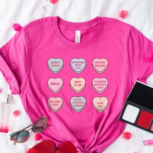 May include: Pink t-shirt with a graphic of nine candy hearts with different sayings on them. The sayings include "Sweet Tooth", "Abscessed With You", "Ahhhh-Mazing", "Smile", "Mint To Be", "Flossed Without You", "Filling You", "Hit Bi-Cuspid Arrow", and "I Pick You".
