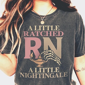 May include: Dark gray t-shirt with the text "A Little Ratched RN A Little Nightingale." The design includes a pink and tan RN, a skeleton hand holding a lamp, and text in a vintage style. The shirt is a crew neck.