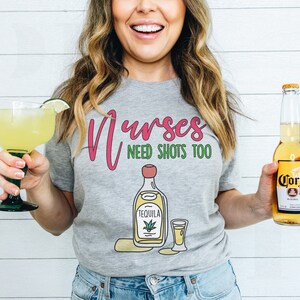 May include: A gray t-shirt with the text "Nurses Need Shots Too" in pink and green. The shirt features a cartoon illustration of a bottle of tequila and a shot glass.