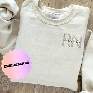 May include: A beige sweatshirt with the letters "RN" embroidered in pink and white flowers. The name "Sarah" is embroidered in black script inside the "R". The sweatshirt has a tag that says "EMBROIDERED" on it.