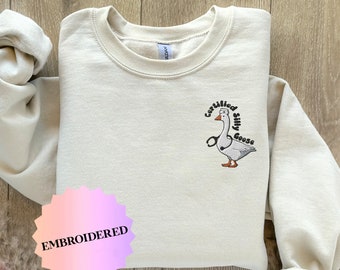 May include: A beige sweatshirt with an embroidered design of a goose wearing a nurse's hat and holding a stethoscope. The text around the goose reads "Certified Silly Goose". The sweatshirt has a pink and white starburst sticker that says "Embroidered" on the bottom left corner.
