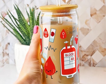 May include: A clear glass reusable tumbler with a bamboo lid and a straw. The tumbler has a graphic design of blood drops, a blood bag, and the text "Give Blood".