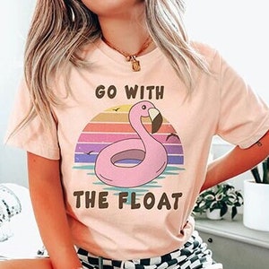 May include: A pink t-shirt with a graphic of a flamingo on a pink inflatable ring in front of a sunset. The text "Go with the float" is printed on the shirt.