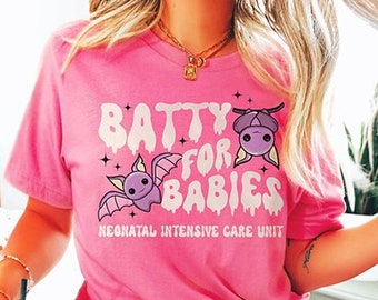 以下が含まれることがあります： 白いグラフィックが「Batty for Babies」と書いてあるピンクのTシャツ。文字は垂れ下がったフォントで、漫画の蝙蝠が2匹描かれている。グラフィックの下のテキストは「Neonatal Intensive Care Unit」と書いてある。