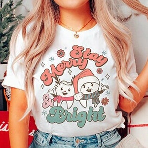 May include: White t-shirt with a Christmas themed graphic featuring two cartoon teeth wearing Santa hats and reindeer antlers. The text "Stay Merry & Bright" is written in a retro font.