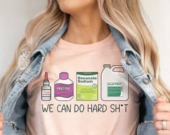 Może przedstawiać: Jasnoróżowa koszulka z napisem "WE CAN DO HARD SH*T" i ilustracjami artykułów medycznych. Koszulka jest zawiązana w talii i noszona z dżinsową kurtką i szarymi spodniami dresowymi.