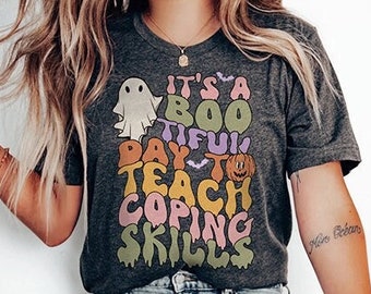 Puede incluir: Una camiseta gris con un gráfico colorido que dice "It's a Boo-tiful Day to Teach Coping Skills". El gráfico incluye un fantasma, una calabaza y murciélagos. La camiseta la lleva puesta una persona con el pelo largo rubio y unos vaqueros azules.
