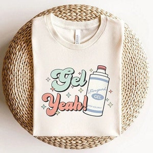 May include: A white t-shirt with a graphic design of a bottle of gel with the text "Gel Yeah!" in a retro font. The bottle is blue and pink with the text "Ultrasound Gel" on it.
