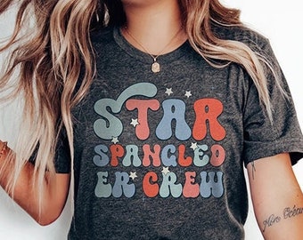 May include: A dark gray t-shirt with a retro-style graphic that says "Star Spangled Banner Crew" in colorful letters with stars. The shirt is being worn by a person with long blonde hair and blue jeans.