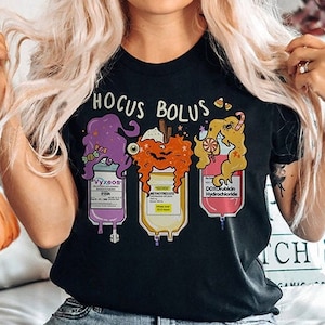May include: Black t-shirt featuring the words "Hocus Bolus" and three cartoon IV bags. The bags are purple, orange, and pink, each with a Halloween design. The shirt is worn by a person.