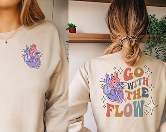 May include: Two women wearing beige sweatshirts with different graphic designs. One sweatshirt features a stylized anatomical heart with the text "Go with the Flow" in rainbow colors. The other sweatshirt features a stylized anatomical heart.
