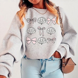 May include: A light grey sweatshirt with a design of pink bows and brain illustrations. The sweatshirt has a crew neck and long sleeves. The model is wearing light blue jeans and a small brown purse.