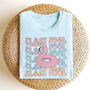 May include: Light blue t-shirt featuring a pink flamingo pool float graphic and the text "FLOAT POOL" repeated in a rainbow colour scheme.
