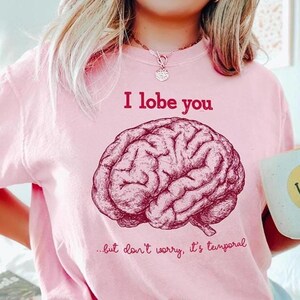 May include: Pale pink t-shirt featuring the words "I lobe you" above a brain illustration. The text "...but don't worry, it's temporal" is below the brain. A white mug with a yellow smiley face is held in the hand.