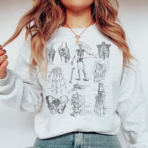 May include: A grey sweatshirt featuring a vintage anatomical illustration of a skeleton, showcasing various bones and joints. The illustration is in black and white and includes the skull, rib cage, pelvis, and limbs.