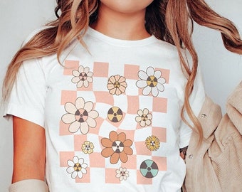 May include: White t-shirt with a pink and white checkered pattern featuring a design of stylized flowers with a nuclear symbol in the center.