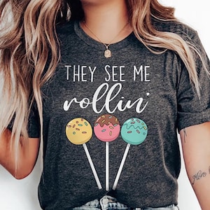 May include: Dark gray t-shirt with the phrase "THEY SEE ME rollin'" in white script. Below the text are three colorful lollipops on sticks: yellow, pink, and teal. The lollipops have sprinkles and chocolate drizzle.
