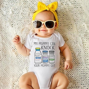 May include: A white baby onesie with the text "My Mommy Can Knock Your Mommy Out" and a graphic of three vials labeled "Propofol", "Precedex", and "Etomidate". The baby is wearing yellow sunglasses and a yellow headband.
