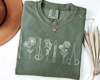 May include: A green t-shirt with a white graphic design of flowers and medical tools. The text "COMFORT COLORS" is visible on the tag.