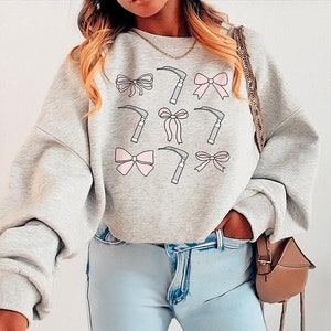 May include: A gray sweatshirt with a graphic design of pink bows and medical tools. The sweatshirt has a relaxed fit and a crew neck.
