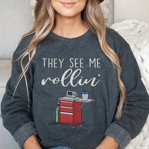 May include: A dark gray sweatshirt with the text "They See Me Rollin'" in white. A cartoon image of a medical cart with a medical device and a water bottle is below the text.