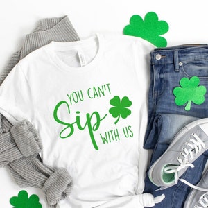May include: White t-shirt with green shamrock and the text "You Can't Sip With Us". The shirt is laid out on a white surface with a gray sweater, blue jeans, and green shamrock decorations.