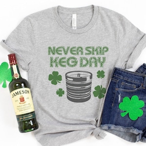 May include: A gray t-shirt with the text "Never Skip Keg Day" and a green shamrock design. A bottle of Jameson Irish Whiskey and a pair of blue denim shorts with a green shamrock patch are also pictured.
