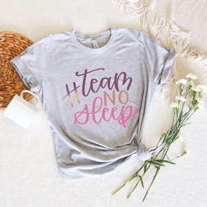 May include: A light gray t-shirt with the text "#Team No Sleep" in a gradient of purple, orange, and pink. The shirt is knotted at the bottom and is displayed on a white surface with a white mug and flowers.