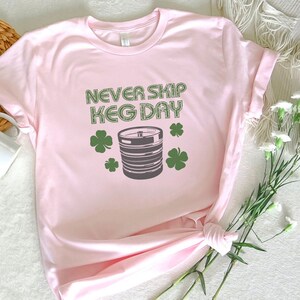 May include: Pink t-shirt with the text "Never Skip Keg Day" in black and green. A green shamrock is on either side of the text. A gray keg with a black tap is in the center of the shirt.