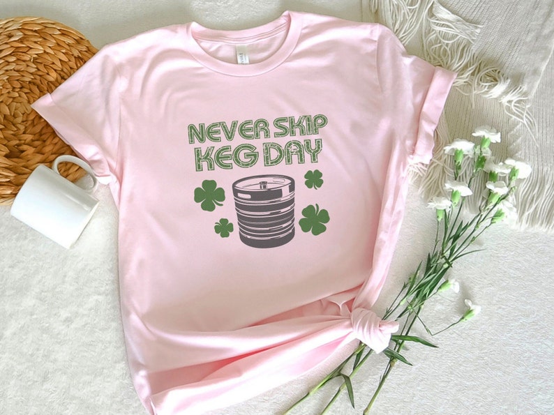 May include: Pink t-shirt with the text "Never Skip Keg Day" in black and green. A green shamrock is on either side of the text. A gray keg with a black tap is in the center of the shirt.